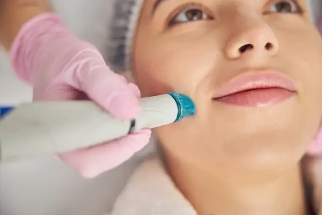 Close-up of woman receiving microdermabrasion facial treatment with handheld device applied to her cheek by aesthetic professional wearing pink gloves