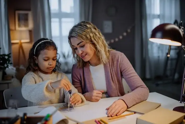 Digital nomad mother helping young daughter with homeschool lessons at home office desk with laptop and educational materials