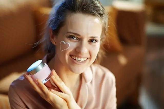 Smiling woman applying heart-shaped skincare cream from pink jar, demonstrating private label skincare product usage