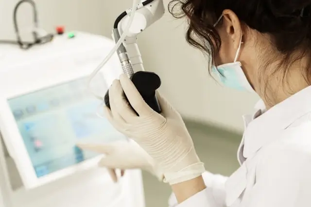 Aesthetic professional in white gloves and surgical mask preparing injection or treatment device in clinical setting with tablet computer visible in background