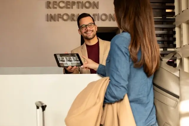 Smiling male receptionist with glasses and beige blazer showing a tablet to a woman in a blue sweater at a modern hotel reception desk