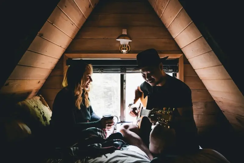Long distance couple bonding in cozy A-frame cabin loft, man playing guitar while woman enjoys coffee during romantic mountain getaway