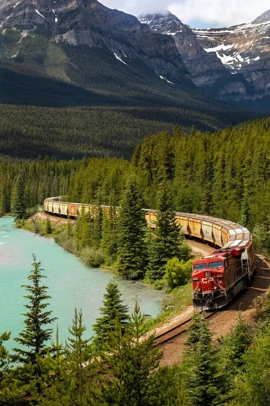 Scenic train journey through Canadian Rockies with red locomotive pulling freight cars along turquoise river surrounded by snow-capped mountains and dense evergreen forest