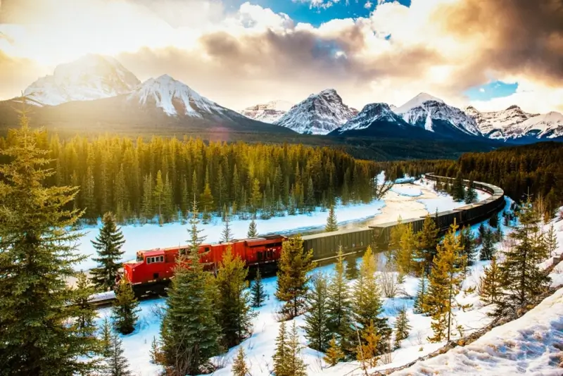 Red passenger train crossing curved bridge through snowy Canadian Rockies landscape with dramatic golden sunset lighting and snow-covered evergreen trees