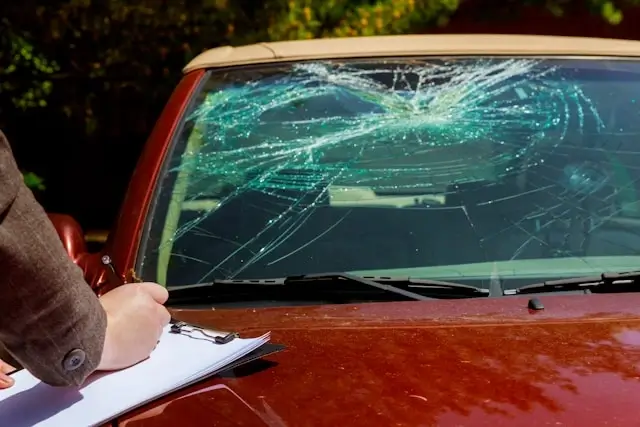 Insurance adjuster documenting cracked windshield damage on red car for collision coverage claim assessment