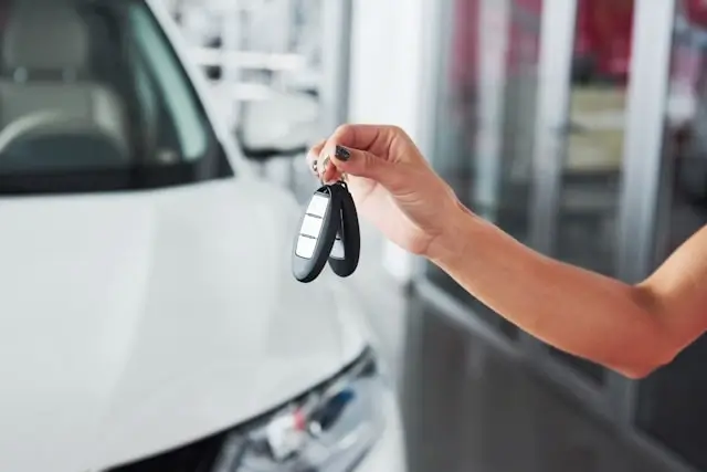 Hand holding car keys next to white vehicle at dealership representing new car purchase requiring car insurance coverage