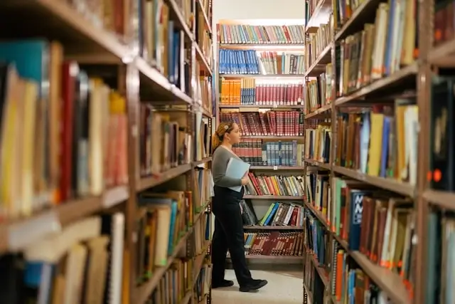 Student researching in library between tall bookshelves filled with academic books for essay writing and study