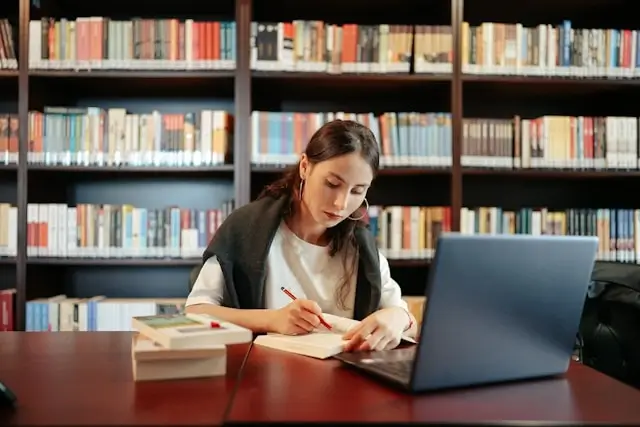 Female student writing essay at library table with laptop and books, demonstrating academic writing process
