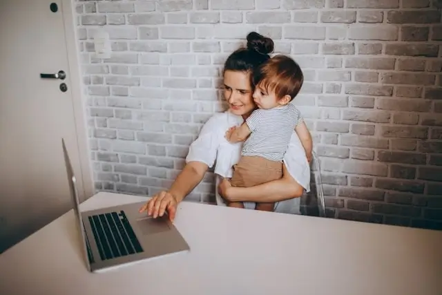Digital nomad parent working remotely on laptop while holding toddler, balancing work and childcare responsibilities at home