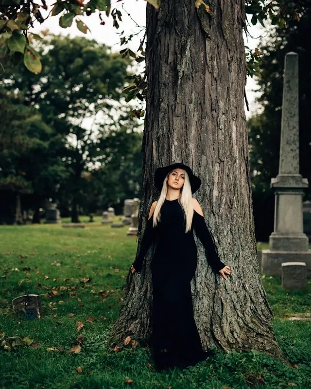 Woman in black long-sleeve maxi dress leaning against large tree in park setting - elegant fall dress ideas for autumn occasions