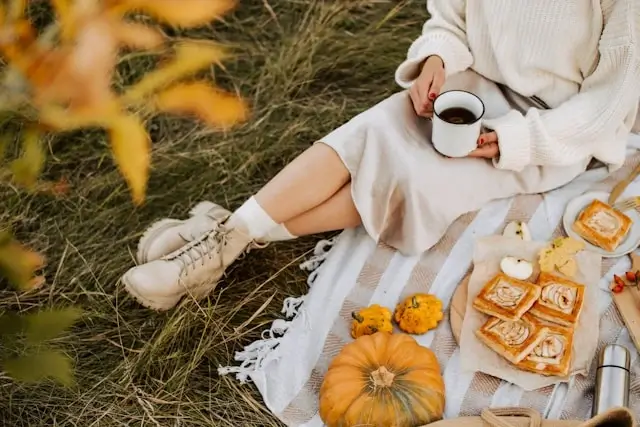 Woman in cream sweater dress enjoying autumn picnic with coffee and pumpkins - cozy fall dress ideas for seasonal outings