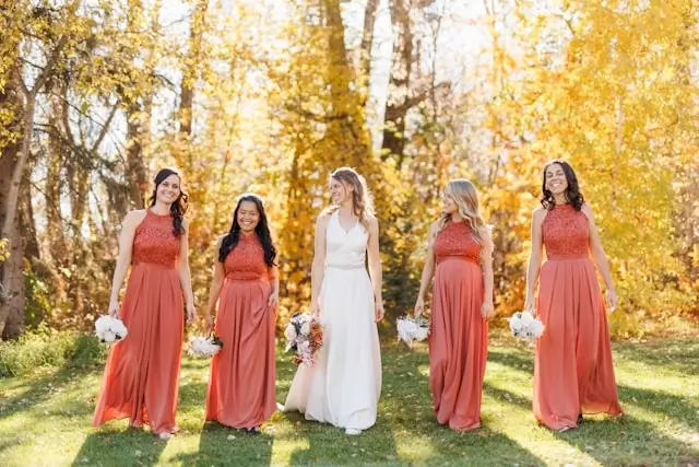 Bride in white gown with bridesmaids wearing coral maxi dresses among golden autumn trees - elegant fall dress ideas for wedding parties