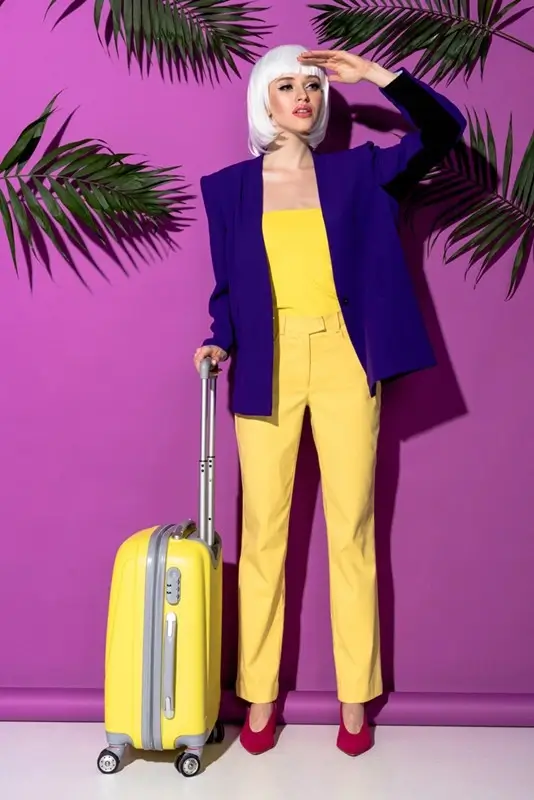 Stylish woman in yellow bodysuit and purple blazer with matching yellow suitcase for colorful travel outfit inspiration
