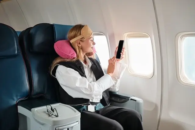 Woman with blonde hair using smartphone while seated in airplane cabin with pink neck pillow and blue leather seats