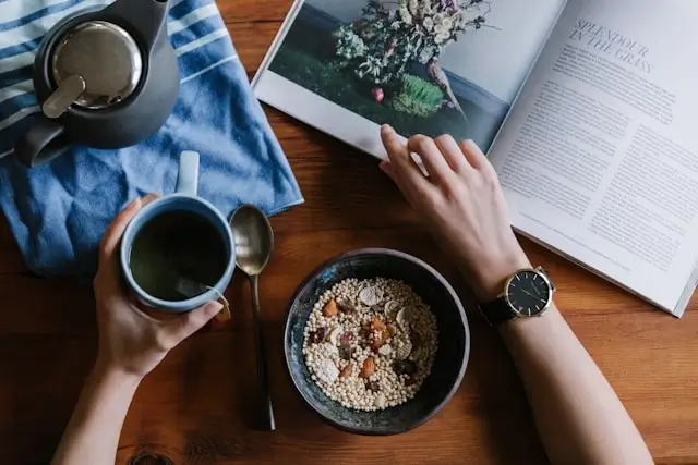 Person enjoying morning routine with coffee, healthy breakfast bowl, and magazine reading at wooden table