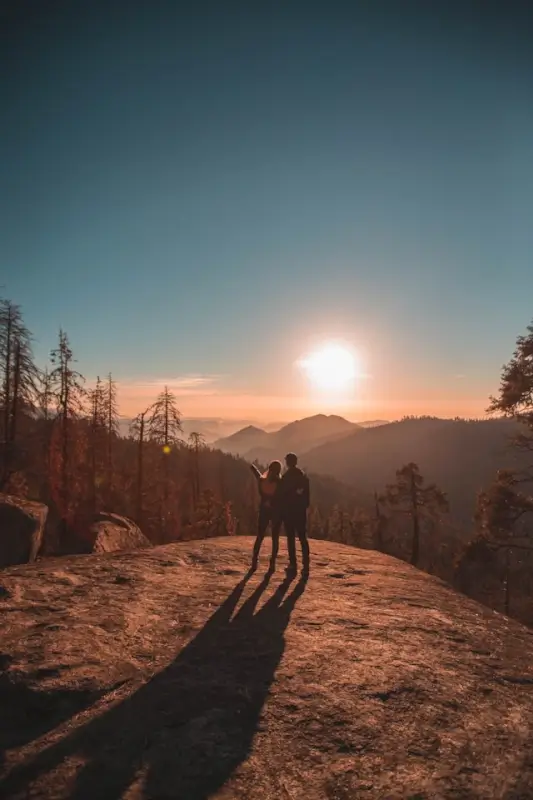 Long distance couple silhouetted against sunset on mountain overlook during hiking adventure, celebrating connection in nature