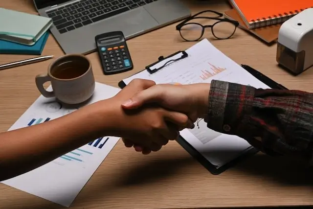 Professional handshake over desk with payroll documents, calculator, and laptop during hiring interview process