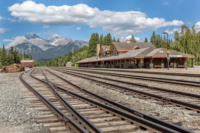 Historic mountain train station with railway tracks and rustic wooden building surrounded by snow-capped Canadian Rocky Mountains under blue sky