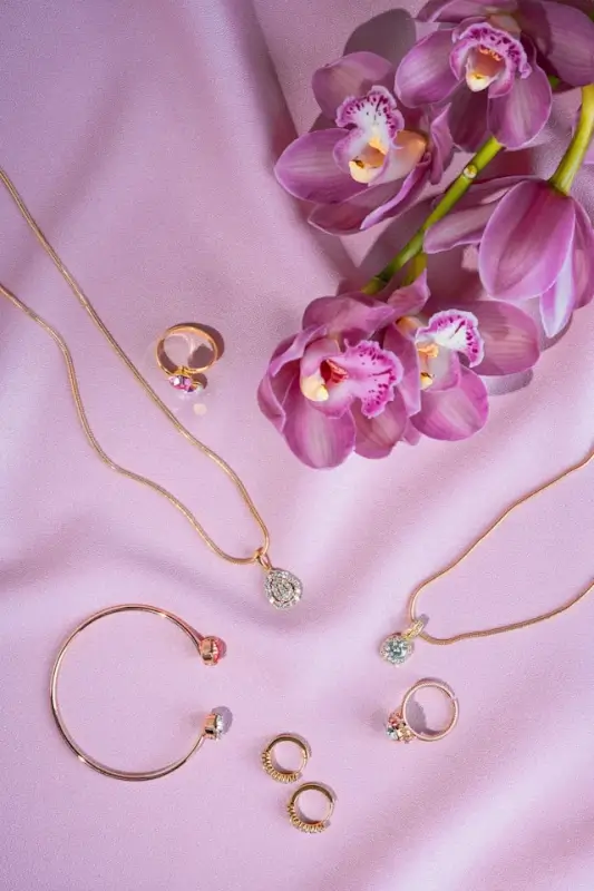 Collection of gold jewelry including necklaces, rings, and bracelet arranged with purple orchid flowers on pink background