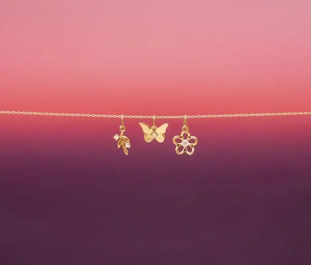 Gold jewelry charms hanging on delicate chain including leaf, butterfly, and flower pendants against pink gradient background