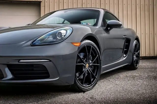 Gray Porsche 718 Cayman sports car with black alloy wheels parked in front of a beige corrugated metal building