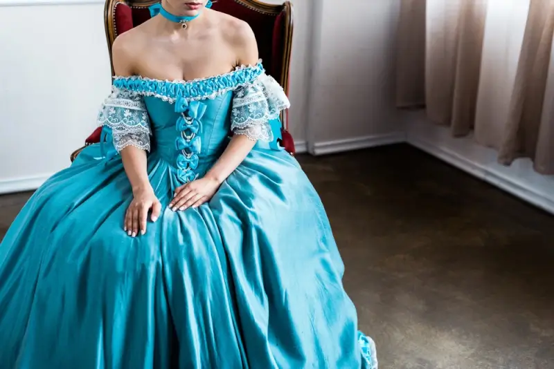 Woman wearing turquoise Renaissance dress with off-shoulder corset bodice, white lace sleeves, and full ball gown skirt while seated in ornate chair