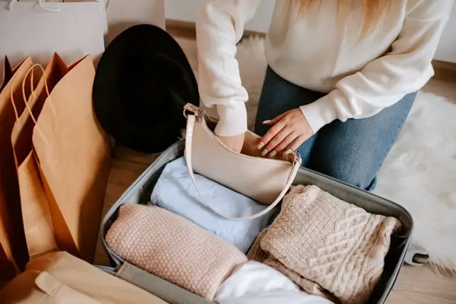 Woman in cream sweater packing folded clothes and accessories into open suitcase, with shopping bags nearby on bed