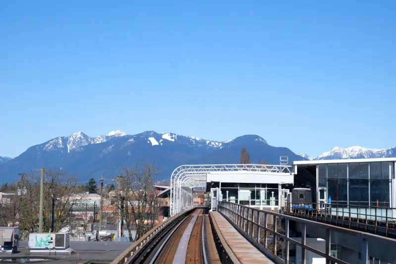 Modern SkyTrain elevated railway track leading toward snow-capped mountains in Vancouver with clear blue sky and urban skyline