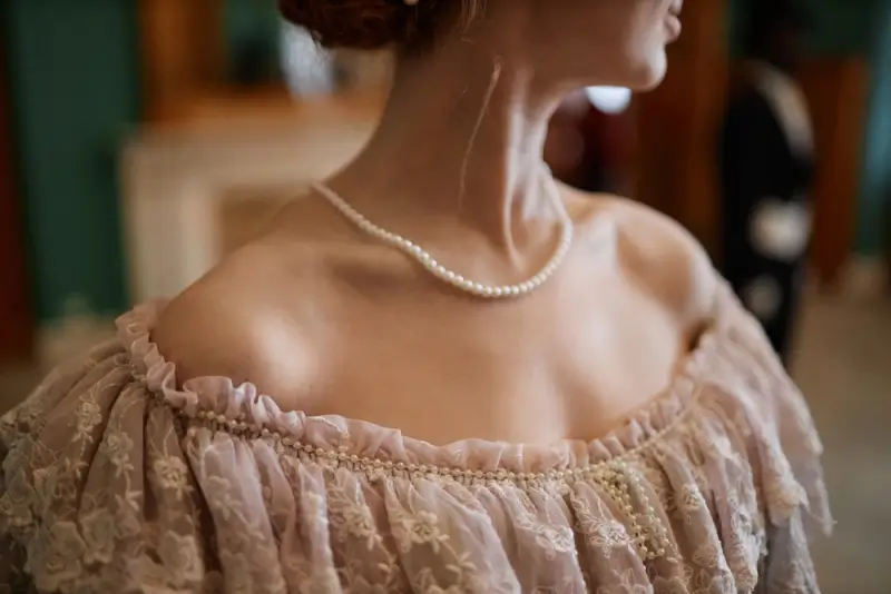 Close-up of woman wearing blush pink Renaissance dress with off-shoulder ruffled neckline, lace details, and pearl necklace accessory