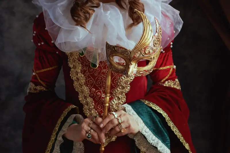 Woman in elaborate red Renaissance dress with gold embroidery and white ruffled sleeves holding ornate Venetian masquerade mask