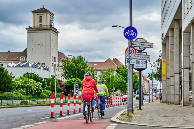 Two cyclists on dedicated bike lane with blue cycling sign and directional signage in German town center