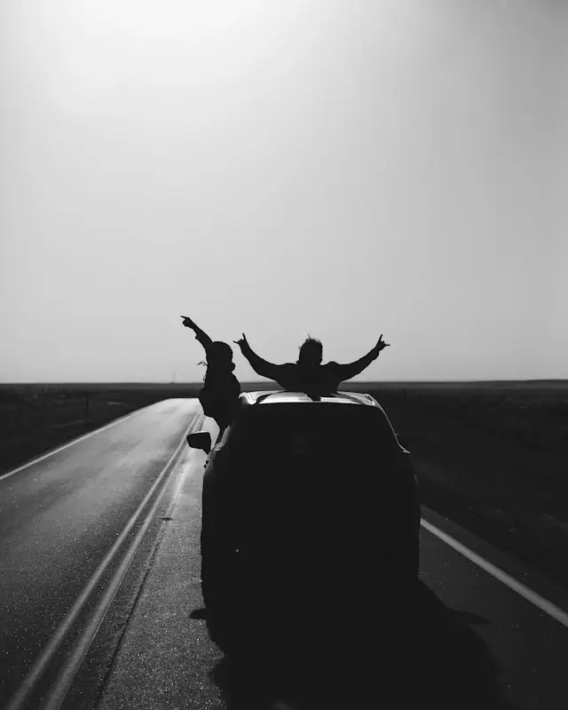 Two people celebrating with arms raised sitting on car roof during road trip on empty highway