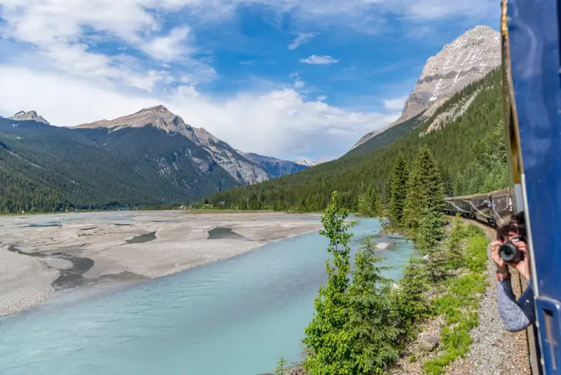 Panoramic view from train window showing turquoise glacial river winding through Canadian Rocky Mountain valley with passengers enjoying scenic railway journey
