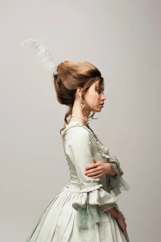 Woman in pale green Renaissance dress with fitted bodice, ruffled details, and period hairstyle with white feather accessory and pearl jewelry