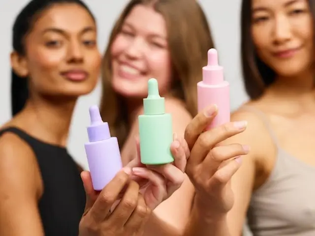 Three diverse women holding colorful private label skincare serums in purple, green, and pink dropper bottles, showcasing product diversity