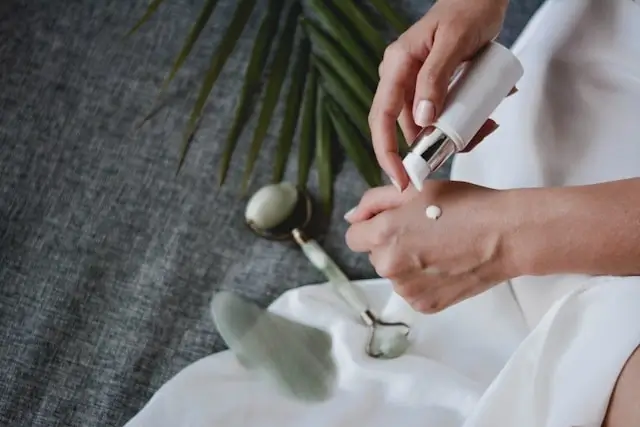 Woman dispensing white private label skincare serum from dropper bottle onto hand with jade roller and palm leaf in spa setting