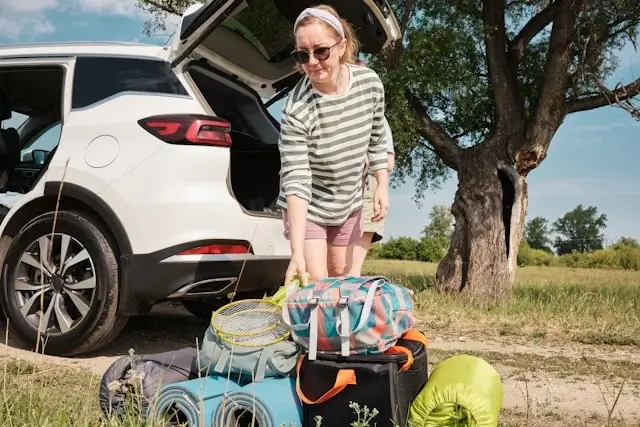 Woman packing travel bags and camping gear from car trunk during road trip preparation