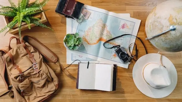 Flat lay of travel planning items on wooden surface including world map, globe, leather backpack, notebook, camera, sun hat, and small potted plant