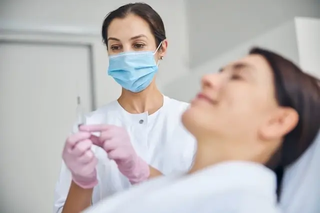 Aesthetic professional in blue surgical mask and pink gloves holding syringe while consulting with female patient in medical office