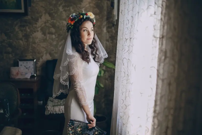 Beautiful Ukrainian woman in traditional wedding dress with floral crown and lace veil in rustic home setting