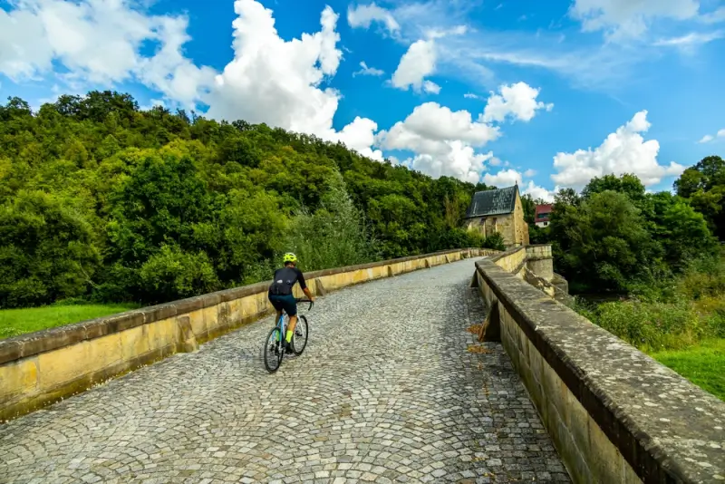 Cyclist riding across historic stone bridge with cobblestone surface through green German countryside