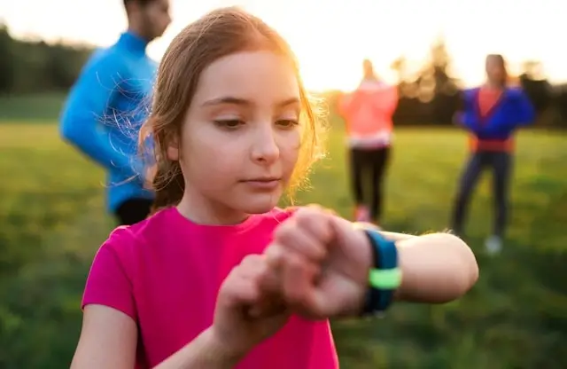 Young girl in pink shirt checking her smartwatch outdoors with family members visible in background during golden hour