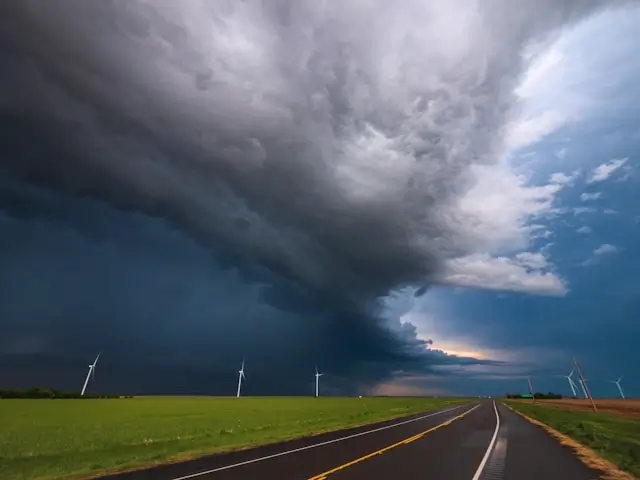 Dramatic storm clouds gathering over empty rural road with wind turbines in distance - showing threatening weather conditions that can suddenly affect travelers