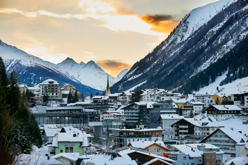 Snow-covered Austrian Alpine village nestled in a dramatic mountain valley at sunset, with traditional chalets and modern buildings scattered across snowy slopes beneath towering peaks