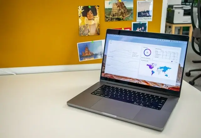 Laptop displaying GPS tracking dashboard with world map and analytics on desk with travel destination photos on wall