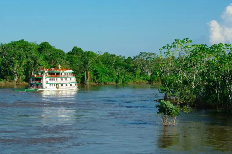 Small white river cruise ship navigating through Amazon rainforest waterway surrounded by dense green jungle vegetation and blue sky