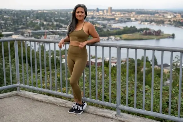 Woman in olive green cotton leggings and matching crop top standing by city overlook railing with lake view