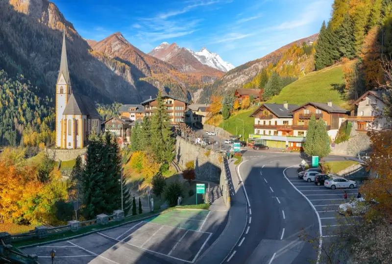 Heilingenblut, the Austrian Alpine village in autumn with distinctive white church spire, traditional wooden buildings, and winding mountain road surrounded by colorful fall foliage and snow-capped peaks