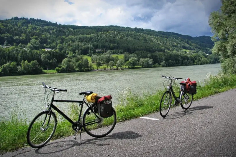 Two touring bicycles with panniers parked on riverside cycling path overlooking Danube River with lush green hills and forest in European countryside