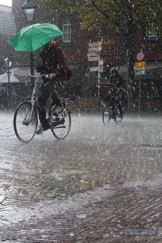 Cyclists riding through heavy rainfall on city street with green umbrella, showing how sudden storms affect bicycle travelers seeking shelter